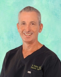 A middle-aged man with short gray hair smiles at the camera, wearing black medical scrubs with Dr. Stephen and Pediatric Dentistry embroidered on the chest, against a teal background.