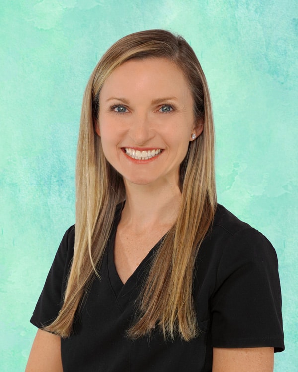 Woman with long, straight blonde hair wearing a black V-neck top, smiling in front of a light teal background.