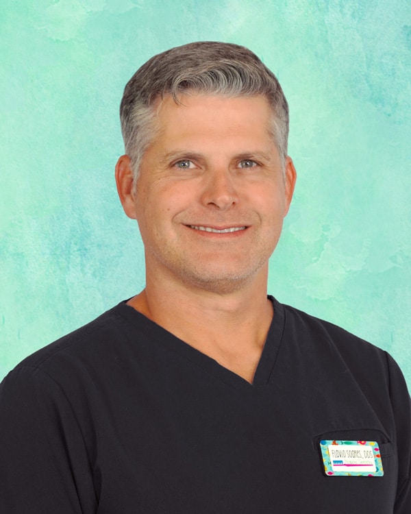 A man with short gray hair, wearing a black V-neck scrub top and a colorful name badge, stands in front of a light blue textured background.