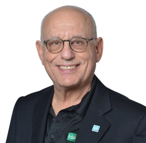 An older man with glasses, wearing a black jacket and shirt with two FDA pins, smiles at the camera against a white background.