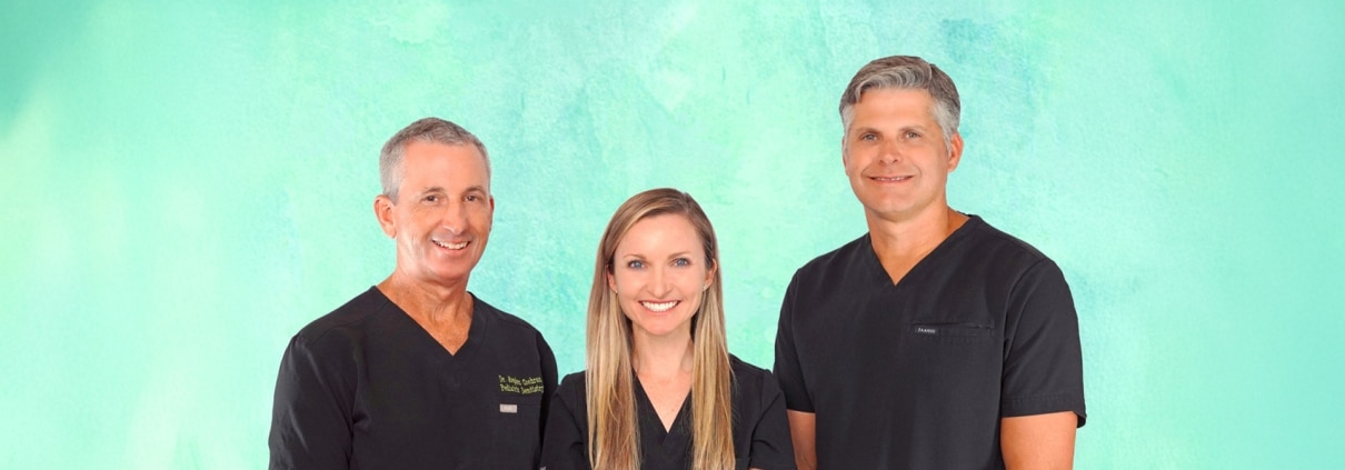Three people wearing black scrubs stand side by side in front of a green background, smiling at the camera.