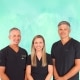 Three people wearing black scrubs stand side by side in front of a green background, smiling at the camera.