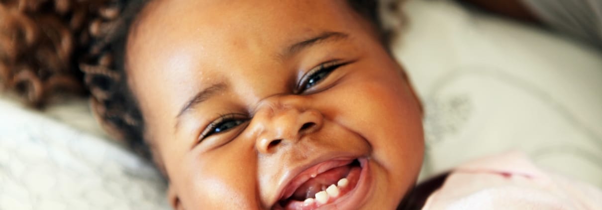A baby lying down and smiling widely, showing several baby teeth, with curly hair and wearing a light pink outfit.