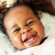 A baby lying down and smiling widely, showing several baby teeth, with curly hair and wearing a light pink outfit.