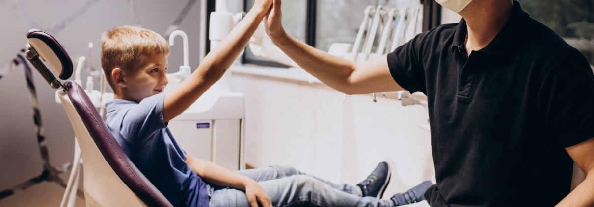 A boy sitting in a dental chair gives a high-five to a masked dentist wearing gloves in a bright dental clinic.