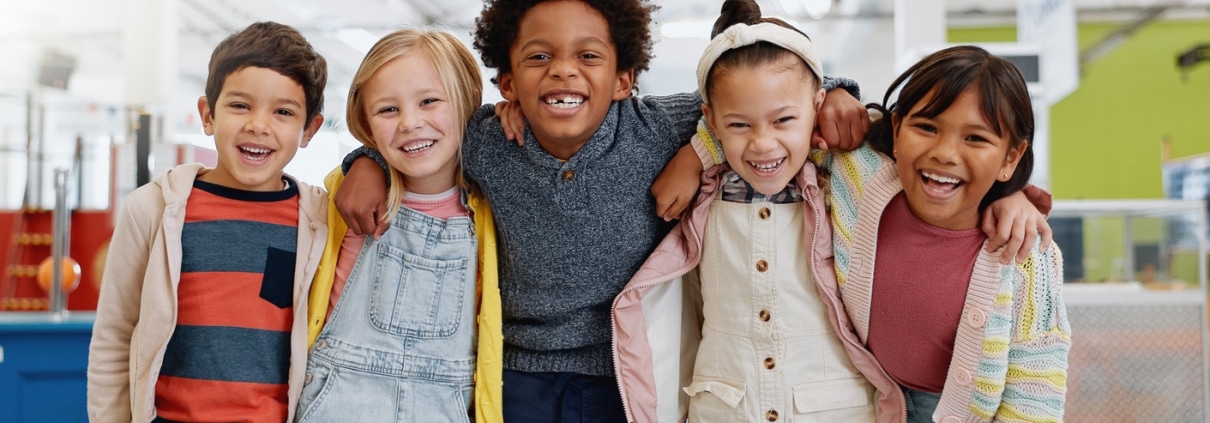 Five children stand indoors with arms around each other, smiling at the camera. They are dressed in casual, colorful clothing. The background shows a bright, open space.