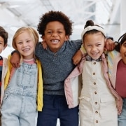 Five children stand indoors with arms around each other, smiling at the camera. They are dressed in casual, colorful clothing. The background shows a bright, open space.