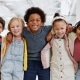 Five children stand indoors with arms around each other, smiling at the camera. They are dressed in casual, colorful clothing. The background shows a bright, open space.